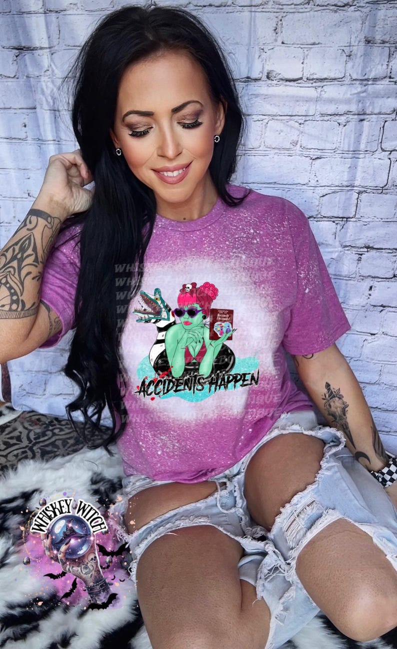 May include: A pink t-shirt with a bleached effect featuring a cartoon illustration of a green monster with a black and white striped creature, sunglasses, and a book that says "Monster Problems". The text "Accidents Happen" is below the illustration.