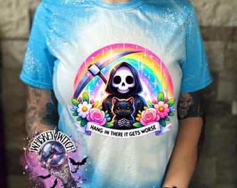 May include: A tie-dye blue t-shirt with a graphic of a cartoon Grim Reaper holding a black cat. The graphic is surrounded by flowers and a rainbow. The text "Hang in there it gets worse" is printed below the graphic.