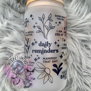 May include: White frosted glass tumbler with a bamboo lid and a straw. The tumbler has black lettering with motivational quotes and illustrations of flowers and leaves. The quotes include "Damn right you will", "I am zen AF", "daily reminders", "Manifest that shit", "You got this baddie", and "Steps today".