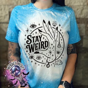 Handmade bleached styles shirt. Close-up of witchy Stay Weird shirt design with celestial artwork, mystical hands, stars, and bold black text