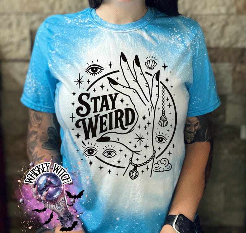Handmade bleached styles shirt. Close-up of witchy Stay Weird shirt design with celestial artwork, mystical hands, stars, and bold black text