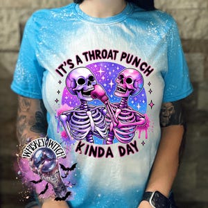 May include: A blue tie-dye t-shirt with a white bleach design featuring two skeletons boxing with the text "It's a throat punch kinda day".
