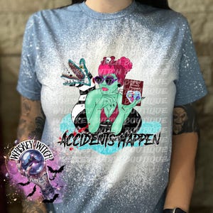 May include: A blue bleached t-shirt with a green and black cartoon character holding a book that says "Recently Deceased". The text "Accidents Happen" is printed on the shirt.