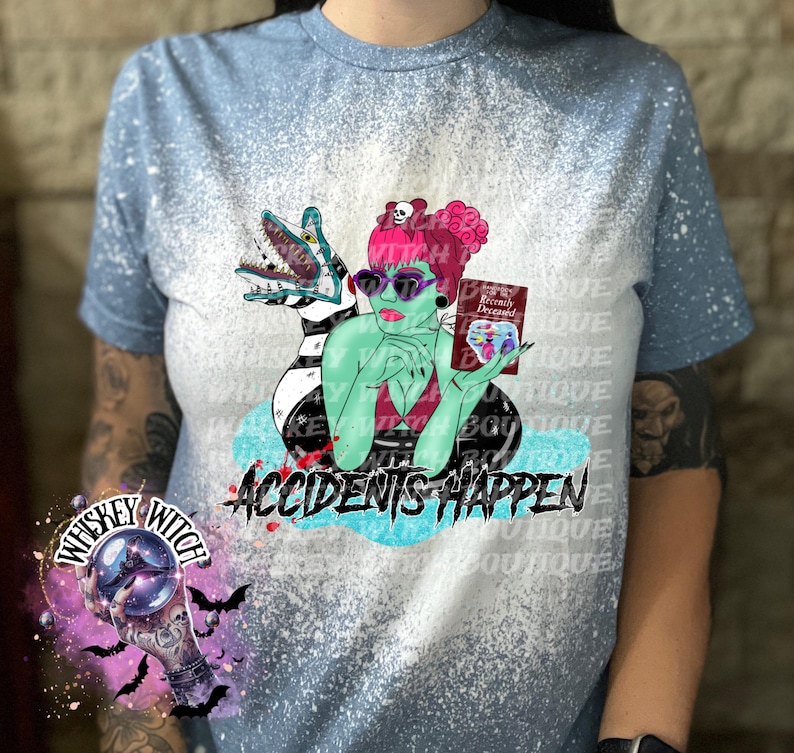 May include: A blue bleached t-shirt with a green and black cartoon character holding a book that says "Recently Deceased". The text "Accidents Happen" is printed on the shirt.