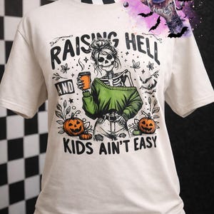 May include: Off-white t-shirt with a skeleton graphic holding a coffee cup, wearing a green sweater and jeans. The text reads "RAISING HELL AND KIDS AIN'T EASY" with pumpkin and bat illustrations. A "Whiskey Witch" logo is visible.