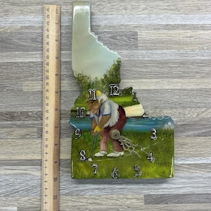 Puede incluir: Un reloj de pared decorativo con la forma del estado de Idaho, con una escena pintada de un golfista. El reloj tiene un paisaje verde y azul con los números del 1 al 12. El reloj mide aproximadamente 30 cm de altura.