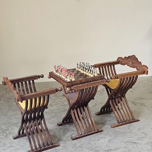 May include: A chess set with a dark wood table and two matching chairs. The chess board is set with red and white pieces. The chairs have a unique curved design with yellow seat cushions. The set is made of dark wood.