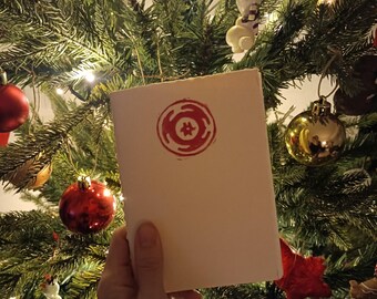 Può includere: Un biglietto bianco con un timbro circolare rosso che presenta un design stilizzato di una stella a quattro punte circondata da un cerchio. Il biglietto è tenuto davanti a un albero di Natale decorato con ornamenti.