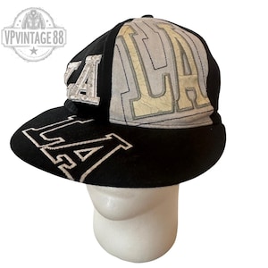 May include: Black and grey baseball cap with "LA" embroidered in white on the brim and side. The top of the cap features "LA" in a distressed, off-white design. The cap is displayed on a white mannequin head.