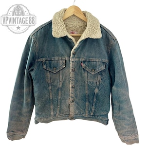 Può includere: Giacca di jeans vintage con fodera in sherpa color crema sul colletto e all'interno. La giacca ha due tasche sul petto, due tasche laterali e una chiusura con bottoni. L'etichetta del marchio Levi's è visibile. La giacca è appesa a una gruccia di legno.
