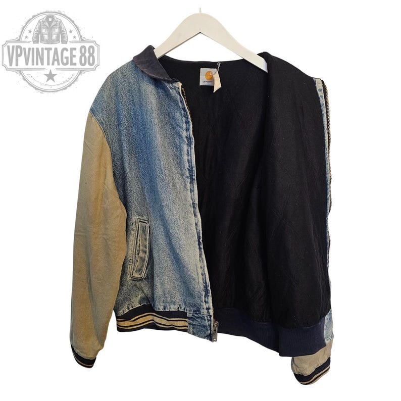 Pu&ograve; includere: Giacca di jeans Carhartt vintage con corpo in denim blu e maniche beige. La giacca ha una fodera interna nera, una chiusura con zip frontale e bordi a coste. La giacca &egrave; appesa a una gruccia bianca.