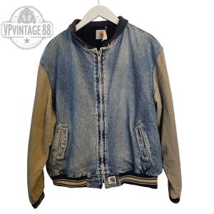 Pu&ograve; includere: Giacca di jeans Carhartt vintage con maniche color cachi e colletto nero. La giacca ha una chiusura a zip, due tasche frontali e orlo a coste. Il logo Carhartt &egrave; visibile sul petto e sull'orlo. La giacca &egrave; appesa a una gruccia bianca.