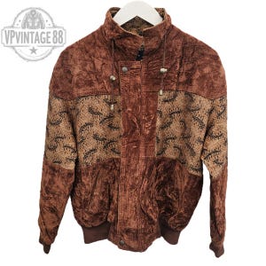 May include: Vintage brown velvet jacket with a high collar and a patterned design on the upper chest and sleeves. The jacket has a button-up front, two pockets, and a ribbed hem. The jacket is hanging on a white hanger.