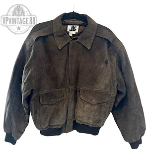 Puede incluir: Chaqueta bomber de ante marrón con cuello, dos bolsillos delanteros y dobladillo y puños de canalé negros. La chaqueta tiene cierre de botones y un pequeño logo en la parte superior del pecho. La chaqueta está colgada de una percha negra.