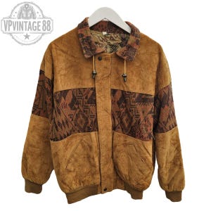May include: A tan suede jacket featuring a brown and black geometric patterned fabric panel across the chest and sleeves. The jacket includes a fold-down collar, a front zip closure with a snap button placket, two front pockets, and ribbed cuffs.