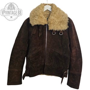May include: A brown suede jacket with a tan faux fur collar. The jacket features a zip closure, two front pockets, and adjustable cuffs. The jacket has a vintage aesthetic. The logo "VP Vintage 88" is in the upper left corner.