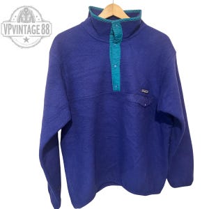 May include: A blue fleece pullover with a teal collar and placket. The pullover has a small chest pocket and a brand logo. The garment is suspended from a wooden hanger.
