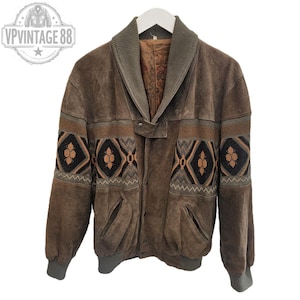 May include: A brown suede jacket featuring a ribbed shawl collar, cuffs, and hem. The garment displays a horizontal band across the chest and sleeves with a black, tan, and brown geometric pattern. It includes two front pockets and a snap closure.