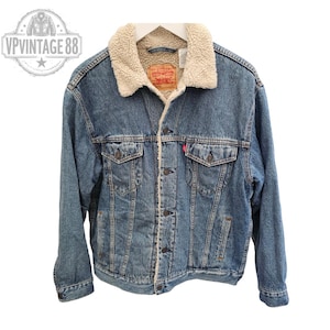 May include: A blue denim jacket with a cream-coloured sherpa collar and lining. The jacket has two chest pockets, two side pockets, and a button-up front. A red Levi's tag is visible on the chest. A classic style.