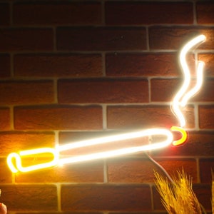 May include: A neon sign shaped like a lit cigarette, emitting a warm, yellow glow against a brick wall. The smoke is depicted with a curved, white neon light. Decorative flowers and wheat stalks are in the foreground.