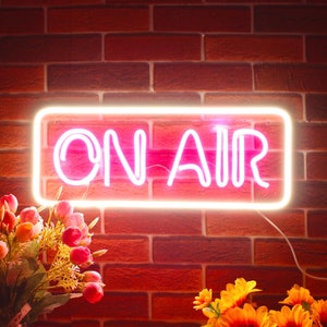May include: A neon sign displaying "ON AIR" in pink, framed by a soft yellow glow. The sign is set against a red brick wall. Floral arrangements, including sunflowers, are visible in the foreground.