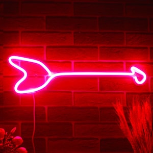 May include: A bright red neon arrow sign against a dark red brick wall. The arrow points to the right, with a heart-shaped fletching. The sign is illuminated, casting a vibrant glow. Decorative flowers and wheat stalks are in the foreground.