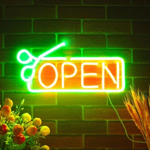 May include: A neon sign with the word "OPEN" in white letters, framed by a green border and scissors graphic. The sign is mounted on a brick wall. Decorative flowers and wheat stalks are in the foreground.