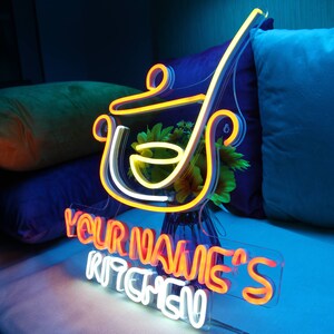 May include: A neon sign in the shape of a cooking pot and ladle, with the text "YOUR NAME'S KITCHEN" in orange and white. The sign is illuminated with yellow and white neon lights, and is placed in front of a vase of flowers.