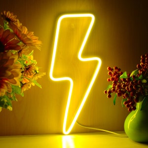 May include: A bright yellow neon lightning bolt sign. The sign is illuminated and has a white cord. The background is a warm yellow color. There are yellow flowers and a green vase with red berries on either side of the sign.