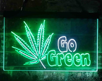 Puede incluir: Un letrero de neón rectangular con una hoja de cannabis verde y las palabras "Go Green" en blanco y verde. El letrero está iluminado con luces de neón brillantes y está montado sobre un fondo oscuro.
