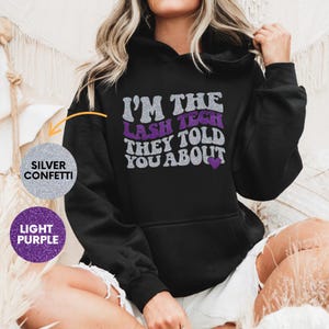 May include: Black hoodie with the text "I'M THE LASH TECH THEY TOLD YOU ABOUT" in silver and purple glitter. The hoodie has a drawstring hood and a front pocket. The text includes a purple heart.
