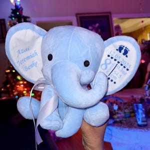 May include: A light blue plush elephant toy with white satin ribbon. The left ear is embroidered with the name Azai Jeremiah Benko. The right ear features footprints, the date 12.05.2025, an infinity symbol, 10.3oz, and 54.5 cm in blue text.