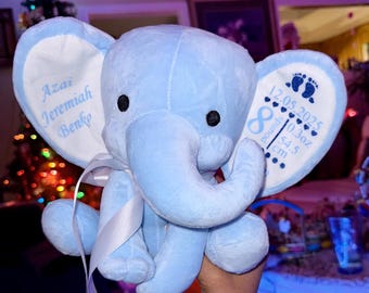 Puede incluir: Un juguete de peluche de elefante azul claro con cinta de satén blanca. La oreja izquierda está bordada con el nombre Azai Jeremiah Benko. La oreja derecha muestra huellas, la fecha 12.05.2025, un símbolo de infinito, 10.3oz y 54.5 cm en azul.