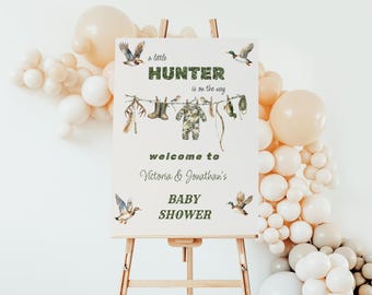 May include: A white sign with green text announcing a baby shower for Victoria & Jonathan. The sign reads "A little Hunter is on the way" and "Welcome to Victoria & Jonathan's Baby Shower". The sign is decorated with a camouflage baby onesie, ducks, and other hunting-themed items.