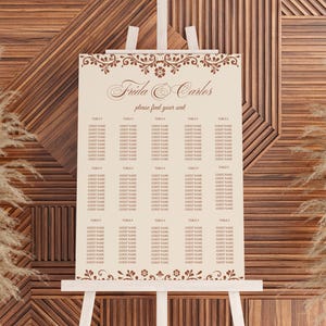 May include: A cream-colored wedding seating chart on a white easel. The chart features the names "Frida & Carlos" and "please find your seat" in elegant script. Floral and scroll designs frame the top and bottom.