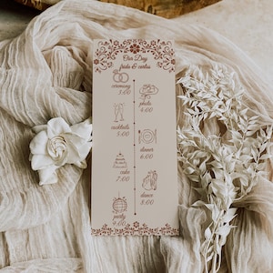 May include: A wedding day timeline card with a floral design in brown. The card lists the schedule for the day, including the ceremony, photo session, cocktails, dinner, cake, dance, and party. A white flower and decorative sprig are in the background.