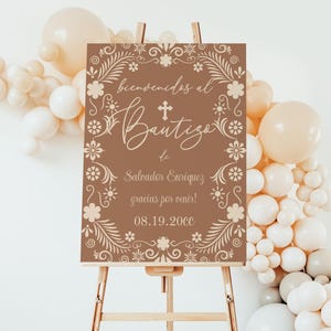 Könnte beinhalten: Ein braunes Schild auf einer hölzernen Staffelei mit den Worten "Bienvenidos al Bautizo" und anderem Text in einer dekorativen Schriftart. Das Schild ist von einem floralen Rand und einem Kreuz umgeben. Ballons in Weiß- und Beigetönen sind im Hintergrund.