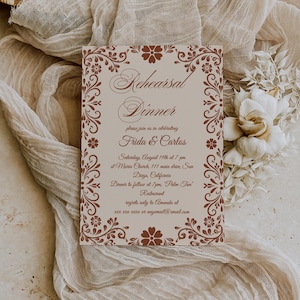 May include: A beige invitation card with brown floral border and text for a rehearsal dinner. The card reads "Rehearsal Dinner" and invites guests to celebrate Frida & Carlos. The card is on a beige cloth with white flowers.