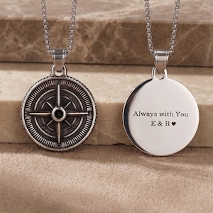 May include: Two silver necklaces. One pendant has a compass design, the other a circular pendant with the engraved text "Always with You E & R ♥". Both necklaces have delicate chain links and are displayed on a textured surface.