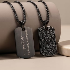 May include: Two black dog tag necklaces. One tag has the inscription "you & me To Eternity and Beyond" in white script. The other tag is covered in small black stones. Both tags hang from a black chain.