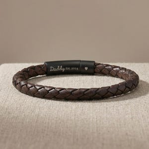 May include: A dark brown braided leather bracelet. The bracelet features a black clasp engraved with the word "Daddy", "Est. 2024", and a heart symbol. The bracelet is displayed on a neutral-toned surface.