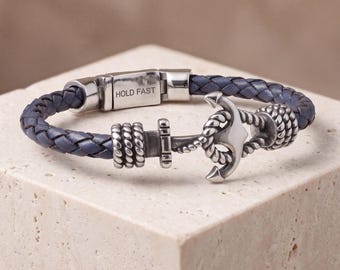 May include: A braided navy blue leather bracelet featuring a silver-toned metal anchor charm and a rectangular clasp engraved with the text HOLD FAST. The anchor design includes rope-like detailing on the shank and flukes of the metal piece.
