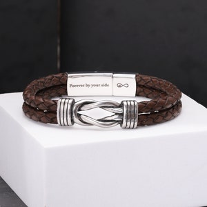 May include: A brown leather bracelet with a braided design. The bracelet has a silver clasp with the inscription "Forever by your side" and an infinity symbol. The bracelet is displayed on a white surface.
