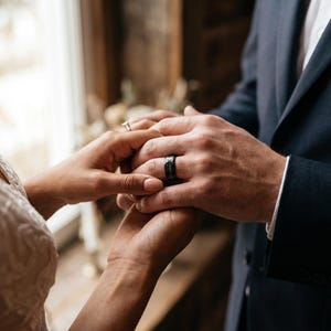 May include: A close-up of two hands, one wearing a black textured ring and the other a delicate engagement ring. The hands are clasped together, with a dark blue suit jacket in the background. The scene suggests a wedding or commitment ceremony.