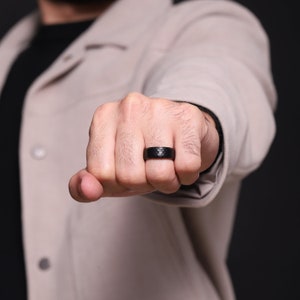 May include: A black ring with a textured surface is displayed on a hand. The ring has a geometric pattern and a dark, matte finish. The background is blurred, with a neutral-toned jacket.