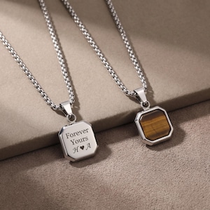 May include: Two silver-toned necklaces with square pendants. One pendant has the engraved text "Forever Yours H♥A". The other pendant features a brown and gold stone. Both necklaces have a chain link design.