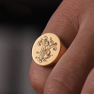 May include: A gold signet ring with a detailed engraved design featuring a stylized letter 'V' surrounded by ornate flourishes.
