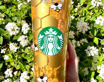 May include: A gold and clear reusable tumbler with a honeycomb pattern and bee illustrations. The tumbler has a green Starbucks logo with a star and a woman's face.