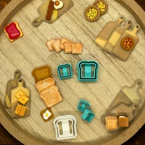 May include: A collection of food-themed cookies and cookie cutters arranged on a wooden tray. The cookies include toast, pizza, crackers, and eggs with bacon. There are also blue and white bread-shaped cookie cutters.
