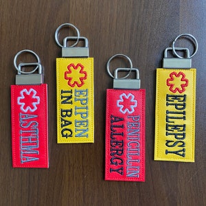 May include: Four yellow and red keychains with a medical symbol and text. The keychains read "EPIPEN IN BAG", "ASTHMA", "PENICILLIN ALLERGY", and "EPILEPSY".